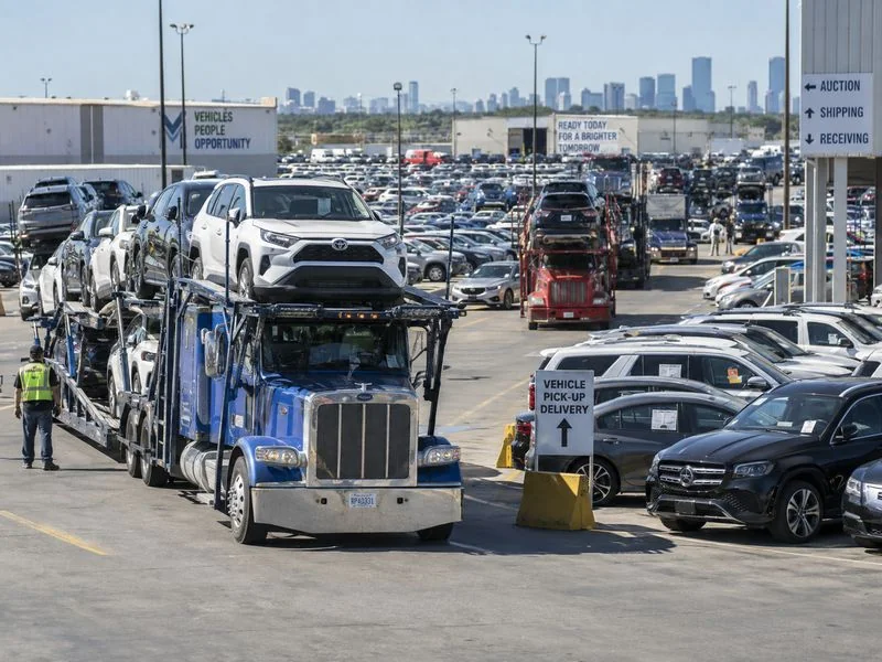 Loaded auto transport truck at a busy vehicle yard during peak shipping season with cars waiting for pickup.